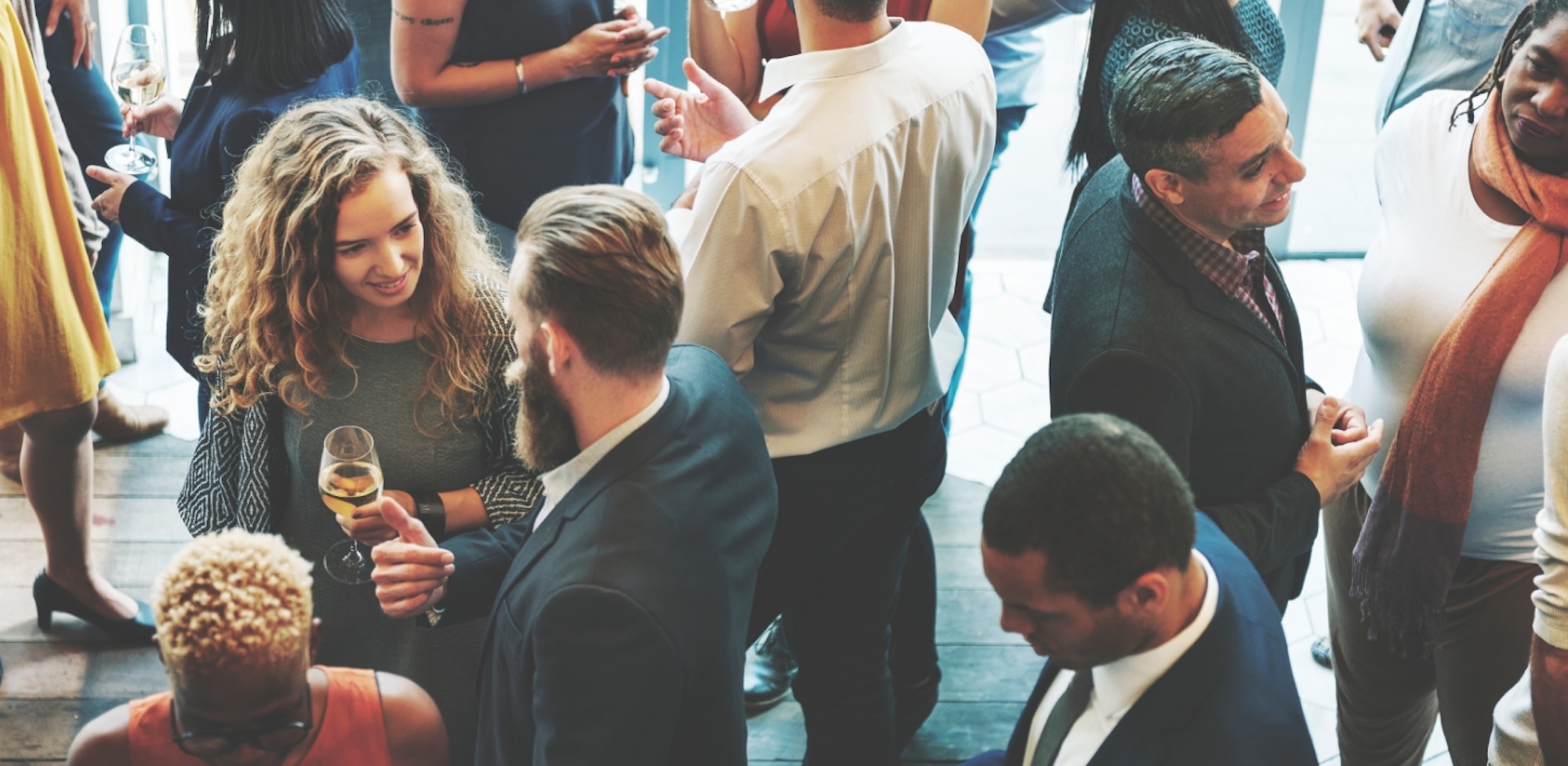 People talking at a reception. A woman with wavey hair chats with a man in a suit, open coloured large beard but we can't see his face